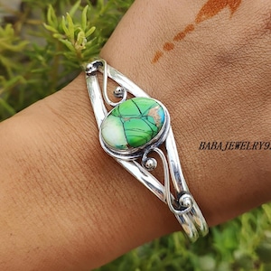 Peut inclure: Un bracelet argenté orné d'une pierre ovale verte et blanche. La pierre présente un motif unique avec des touches de bleu et de marron. Le bracelet ouvert comporte des volutes décoratives et le texte "BABAJewelry93".