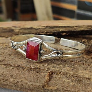 Peut inclure: Un bracelet jonc en argent avec une pierre précieuse rouge sertie dans une bélière en argent. Le bracelet a un design délicat et orné de volutes et de petites perles.