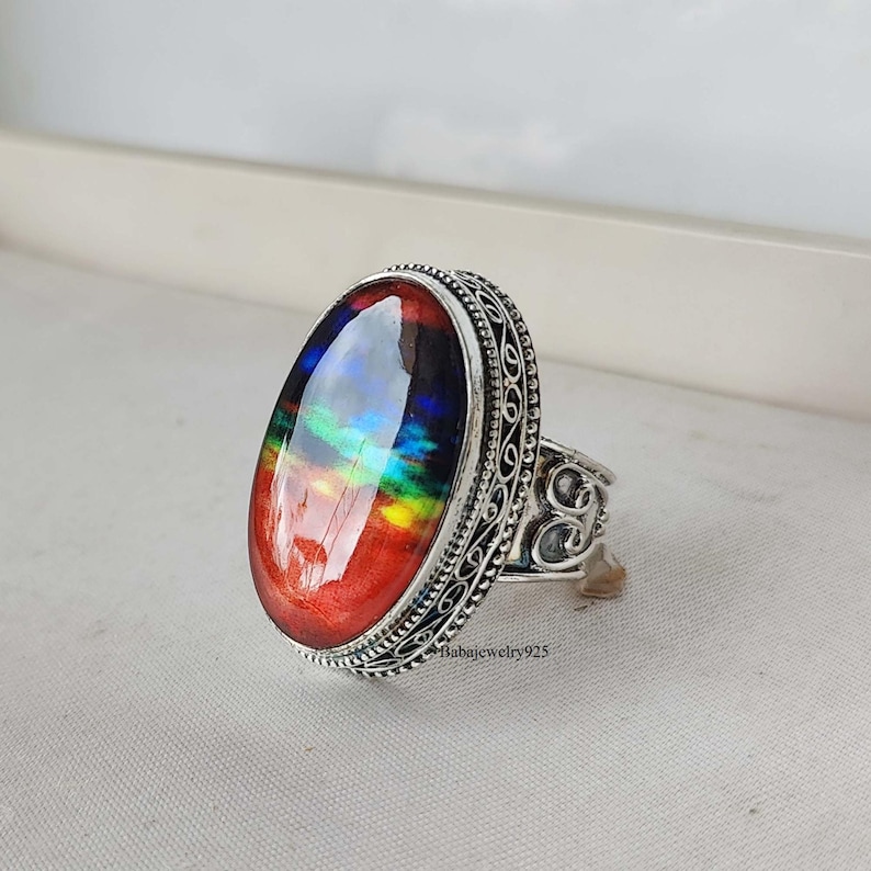 May include: A silver ring with an oval-shaped gemstone. The gemstone is a vibrant red with iridescent rainbow colors. The ring has a filigree design.