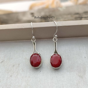 May include: A pair of silver dangle earrings featuring red oval-shaped gemstones. The earrings have a simple design with a curved silver wire.
