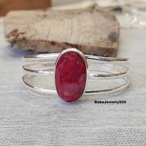 May include: A silver bangle bracelet with a red oval gemstone set in the centre. The bracelet has three bands.