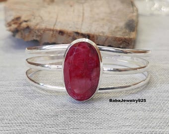 May include: A silver bangle bracelet with a red oval gemstone set in the centre. The bracelet has three bands.