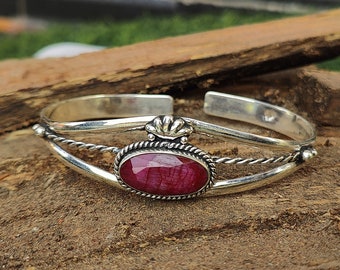 Puede incluir: Un brazalete de plata con una piedra preciosa roja engastada en un bisel de plata. El brazalete tiene un diseño de alambre trenzado y un detalle decorativo en forma de corona.