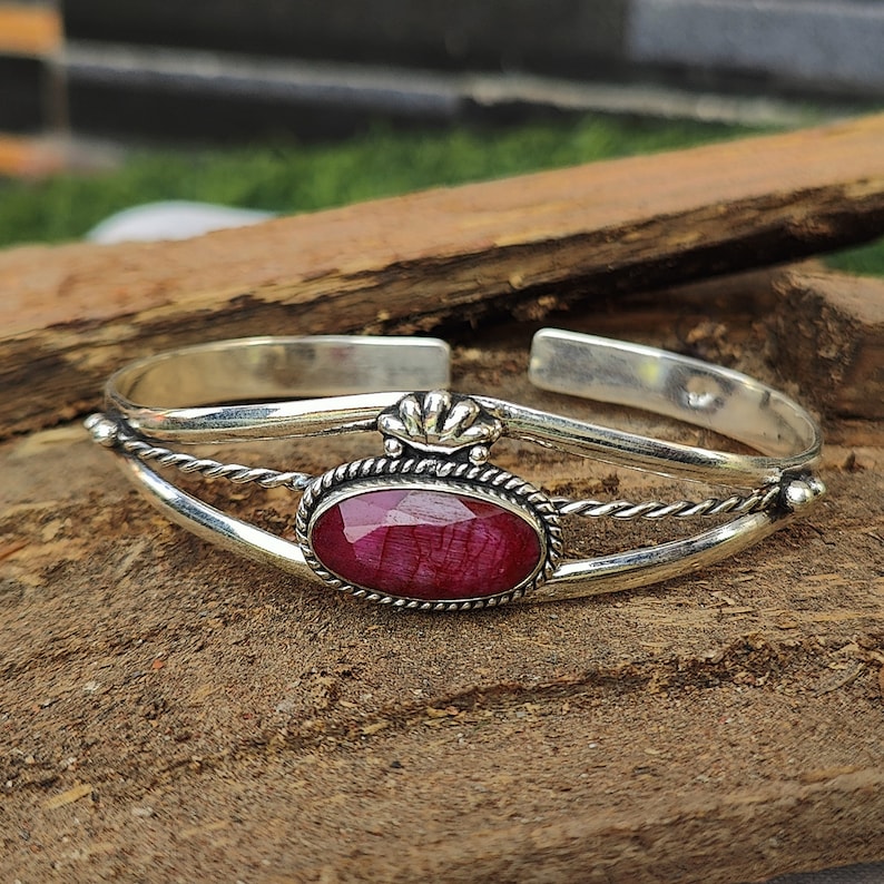 May include: A silver bangle bracelet with a red gemstone set in a silver bezel. The bracelet has a twisted wire design and a decorative crown-like detail.