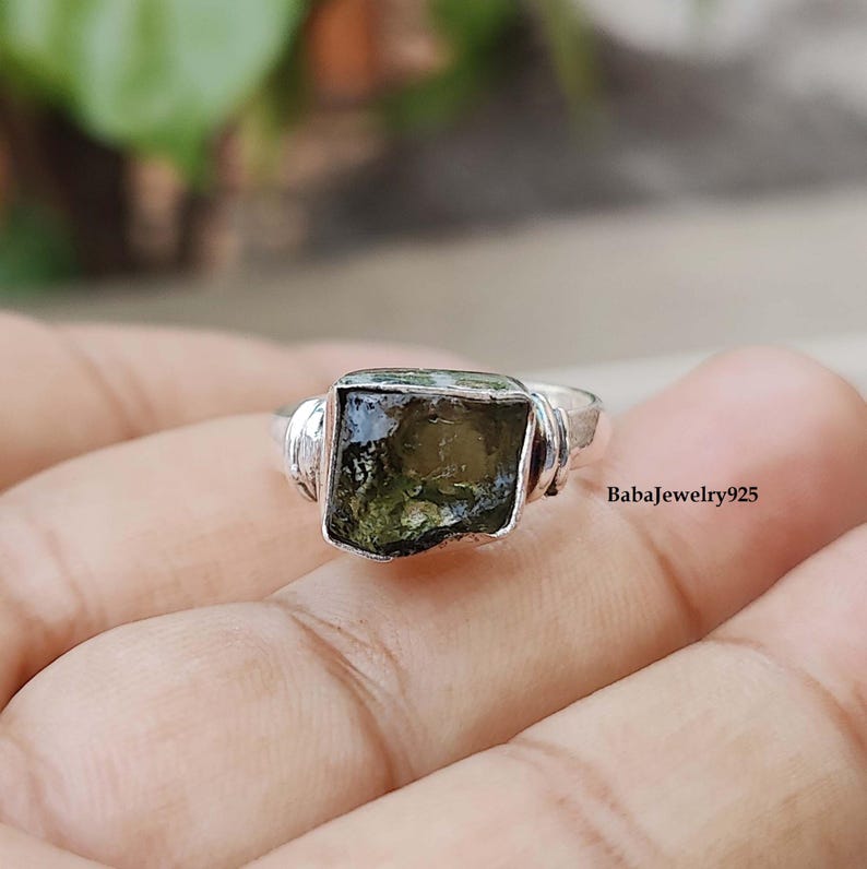 May include: A silver ring with a green gemstone set in a square shape. The ring is set with a bezel setting and has a textured band.
