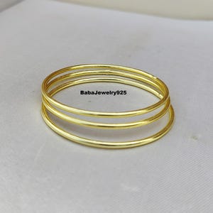 Puede incluir: Tres brazaletes de oro apilados uno encima del otro. Los brazaletes son lisos y brillantes.