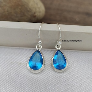 Peut inclure: Une paire de boucles d'oreilles argentées en forme de goutte d'eau avec des pierres précieuses bleu vif. Les pierres sont serties dans une lunette en argent et ont une forme de poire. Le texte "BabaJewelry925" est visible.