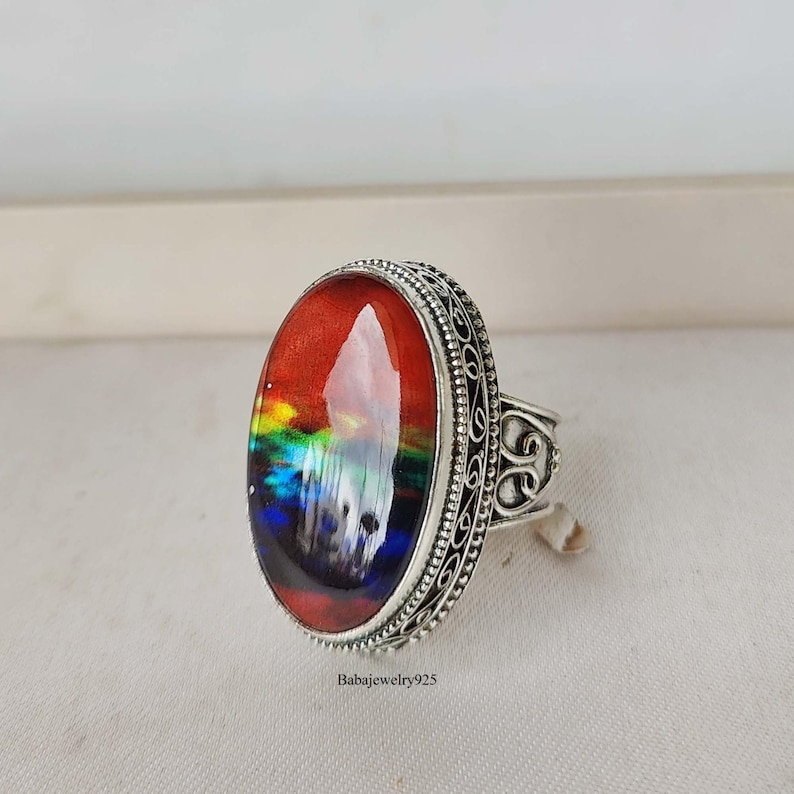 May include: A silver ring with an oval-shaped, multi-colored stone set in a filigree design. The stone has a red, orange, yellow, green, blue, and purple color scheme.