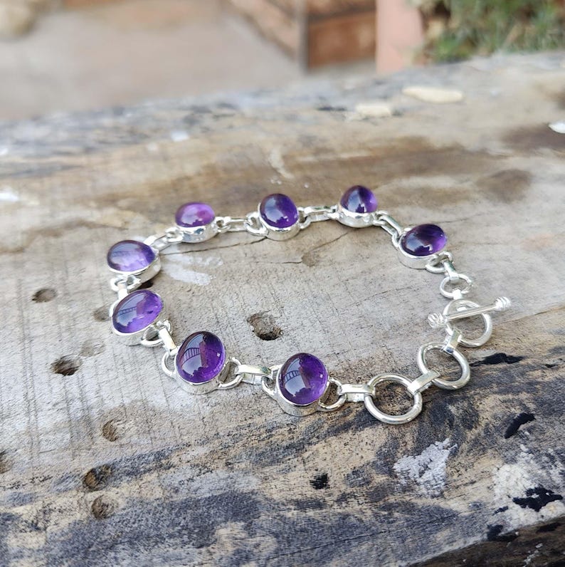 K&ouml;nnte beinhalten: Ein silbernes Kettenarmband mit acht ovalen Amethyst-Edelsteinen. Das Armband hat einen Toggle-Verschluss.