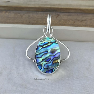 May include: Oval-shaped pendant with a vibrant abalone shell inlay, showcasing iridescent blues, greens, and purples. The shell is set in a silver-toned metal frame with a decorative bail. The pendant is a unique piece of jewelry.