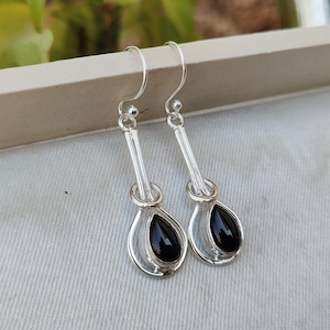 May include: A pair of silver-toned drop earrings with a teardrop-shaped black stone. The earrings have a simple, elegant design with a hook closure. The black stones are smooth and polished, set within a silver frame.
