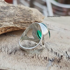 May include: A silver ring with a green gemstone set in a unique, open design. The ring is shown from the side, with the gemstone facing the camera.