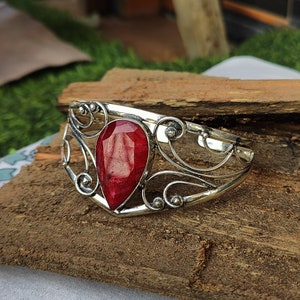May include: A silver bangle bracelet with an ornate design features a large, teardrop-shaped red gemstone.
