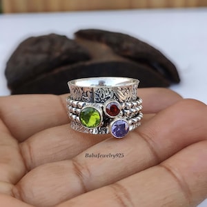 Könnte beinhalten: Ein silberner Drehring mit einem grünen, roten und violetten Edelstein. Der Ring ist mit einem filigranen Blumenmuster verziert.