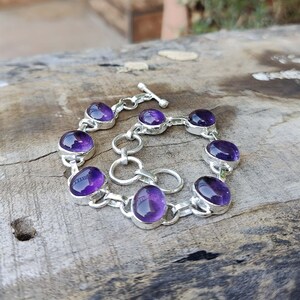 K&ouml;nnte beinhalten: Ein silbernes Kettenarmband mit sieben ovalen Amethyst-Edelsteinen. Das Armband hat einen Schiebeverschluss.