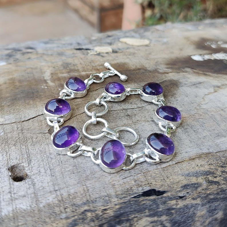 K&ouml;nnte beinhalten: Ein silbernes Kettenarmband mit sieben ovalen Amethyst-Edelsteinen. Das Armband hat einen Schiebeverschluss.