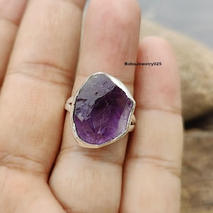 Pode incluir: Um anel de prata com uma grande gema de ametista bruta. A ametista é de cor roxa escura e tem uma forma natural irregular.