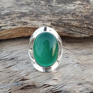 May include: A silver ring with a green oval gemstone set in a decorative silver frame. The ring is on a wooden surface.