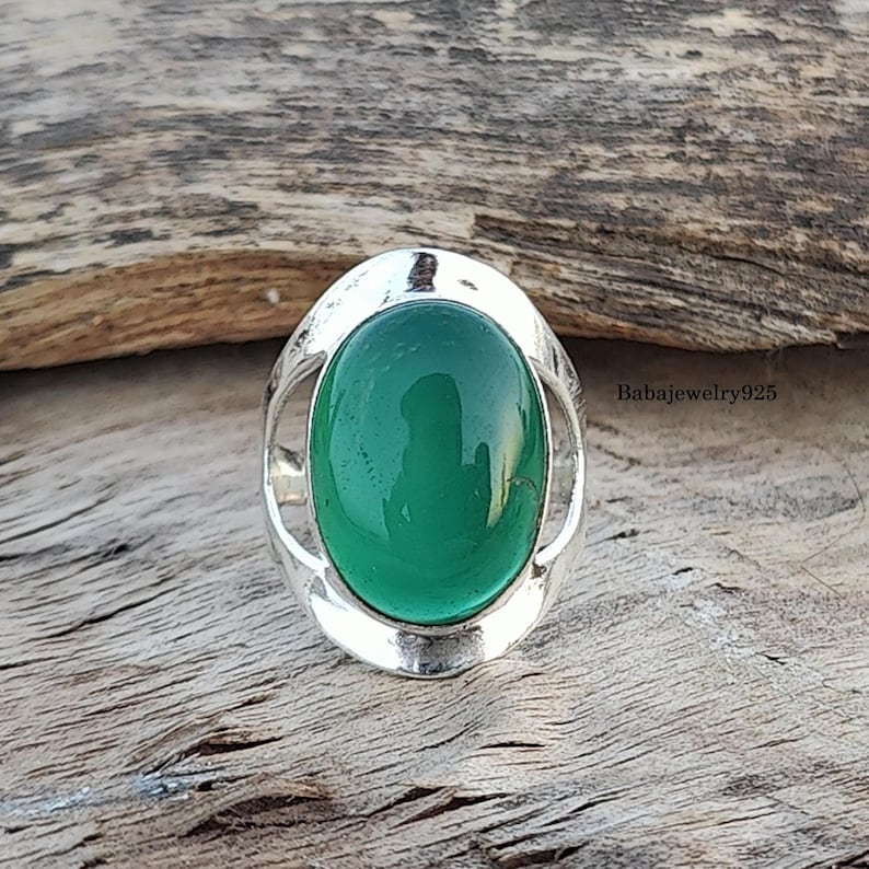 May include: A silver ring with a green oval gemstone set in a decorative silver frame. The ring is on a wooden surface.