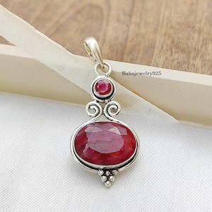 May include: A silver pendant featuring a large, oval-cut ruby gemstone. A smaller, round ruby is set above the oval stone, connected by decorative swirls. The pendant has a bail for a chain and a cluster of small silver beads at the base. The text "Babajewelry925" is visible.