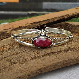May include: A silver bangle bracelet with a red gemstone set in a silver oval. The bracelet has a twisted wire design and a decorative crown-like detail.