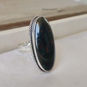 Può includere: Un anello d'argento con una pietra ovale verde e rossa. L'anello ha una fascia intrecciata.
