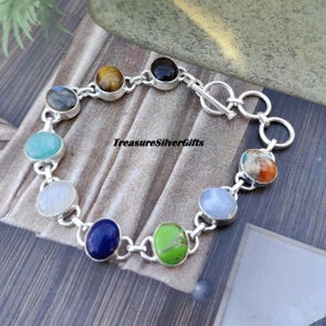 May include: A silver bracelet with a toggle clasp features nine different gemstone cabochons. The stones include amethyst, tiger's eye, black onyx, moonstone, lapis lazuli, turquoise, aventurine, and blue lace agate.  TreasureSilverGifts is printed on the bracelet.