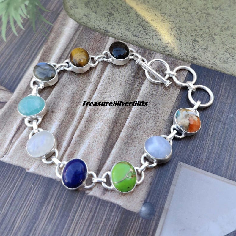 May include: A silver bracelet with a toggle clasp features nine different gemstone cabochons. The stones include amethyst, tiger's eye, black onyx, moonstone, lapis lazuli, turquoise, aventurine, and blue lace agate.  TreasureSilverGifts is printed on the bracelet.