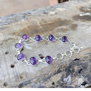 K&ouml;nnte beinhalten: Ein silbernes Kettenarmband mit ovalen Amethyst-Edelsteinen. Das Armband hat einen Schiebeverschluss.