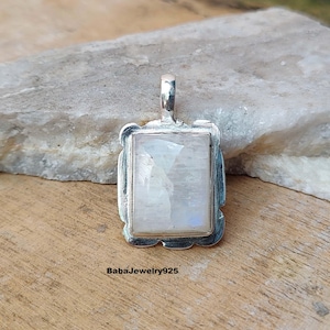 Pode incluir: Um pingente de prata com uma pedra de lua retangular engastada em uma moldura decorativa. O pingente está sobre uma superfície de madeira.