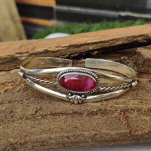 May include: A silver bangle bracelet with a pink oval gemstone set in a silver bezel. The bracelet has a twisted wire design and a decorative accent.