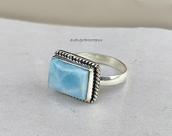 May include: A silver ring featuring a light blue square-shaped gemstone set within a rope-like border.