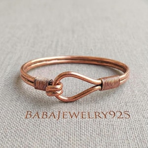 May include: Copper bracelet with a hook and eye clasp. The bracelet is a warm, reddish-brown color and features a looped design for closure. The text "BABAJewelry925" is printed on the surface.