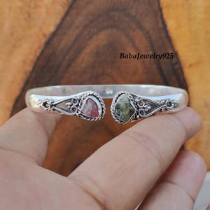 May include: Silver bangle bracelet with two heart-shaped stones. The stone on the left is pink and green, and the stone on the right is green and black. The bracelet has a delicate floral design.