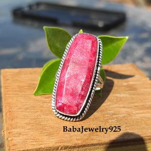 Może przedstawiać: Srebrny pierścionek z dużym, prostokątnym, fasetowanym rubinem. Pierścionek ma prosty, srebrny pasek z wzorem przypominającym sznur.