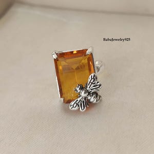 Op de afbeelding: Een zilveren ring met een grote, rechthoekige, amberkleurige edelsteen en een gedetailleerd bijendesign aan de zijkant.