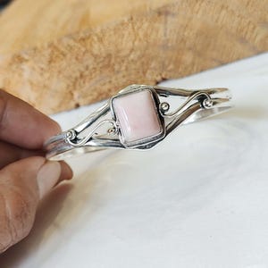 May include: A silver bracelet with a delicate, swirling design and a square pink gemstone in the center.