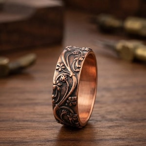 May include: A copper-toned metal ring featuring a raised, embossed floral pattern that wraps around the entire outer band. The design consists of intricate vines, leaves, and small five-petaled flowers with a dark, oxidised finish in the recessed areas.