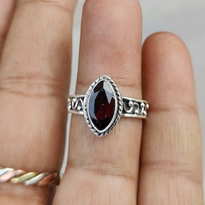 May include: A silver ring with a dark red gemstone set in a marquise shape. The ring has a delicate, ornate design.