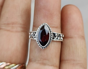 May include: A silver ring with a dark red gemstone set in a marquise shape. The ring has a delicate, ornate design.