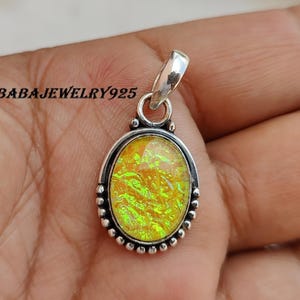 Puede incluir: Colgante ovalado con una fianza y engaste plateados. El colgante presenta una piedra de color amarillo-verdoso vibrante con un aspecto texturizado e iridiscente. El engaste está detallado con pequeños adornos redondos. El texto "BABAJEWELRY925" es visible.