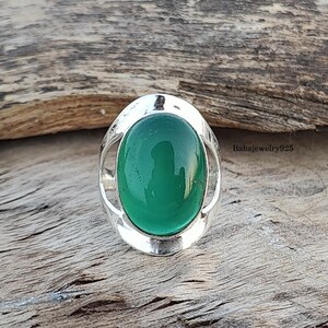 May include: A silver ring with a green oval gemstone set in a decorative silver frame. The ring is on a wooden surface.