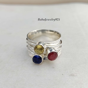 May include: A silver ring with three gemstones: a yellow, a red, and a blue. The ring is made of three bands stacked on top of each other.