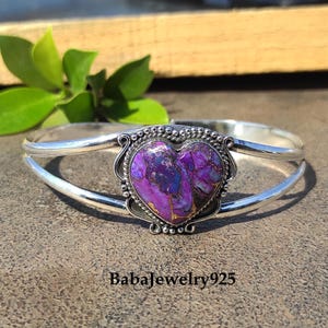 May include: A silver bangle bracelet with a heart-shaped purple and blue stone set in a silver bezel. The stone has a marbled pattern and is surrounded by a decorative silver border.