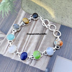 May include: A silver bracelet with a toggle clasp features nine different gemstone cabochons. The stones are set in silver and include labradorite, tiger's eye, aquamarine, moonstone, lapis lazuli, turquoise, and a multi-colored stone.