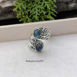 May include: A silver ring featuring two blue labradorite gemstones set in a twisted band with ornate detailing. The ring is displayed on a white surface.