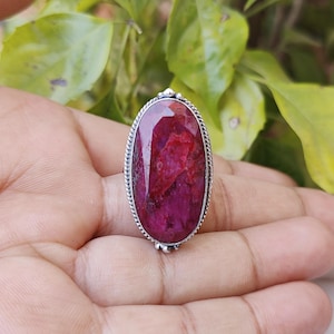 May include: A silver ring with a large, oval-shaped, red gemstone. The gemstone has a deep red color with a marbled pattern.