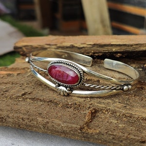 May include: A silver bangle bracelet with a red oval gemstone set in a silver bezel. The bracelet has a twisted wire design and a decorative floral accent.