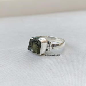 May include: A silver ring with a square green gemstone. The ring is set in a simple band with a textured detail on the side.