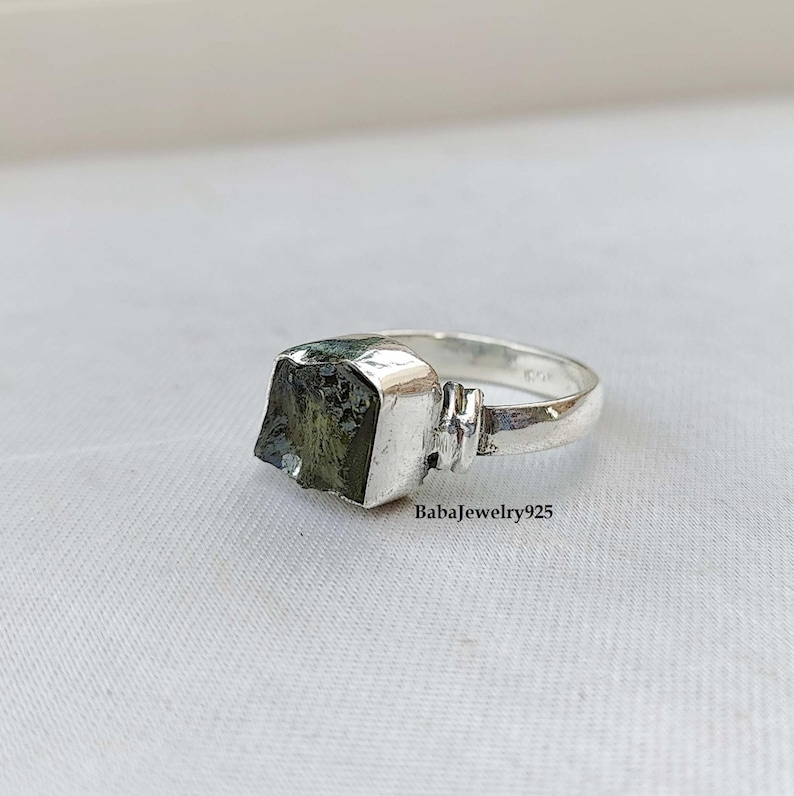 May include: A silver ring with a square green gemstone. The ring is set in a simple band with a textured detail on the side.